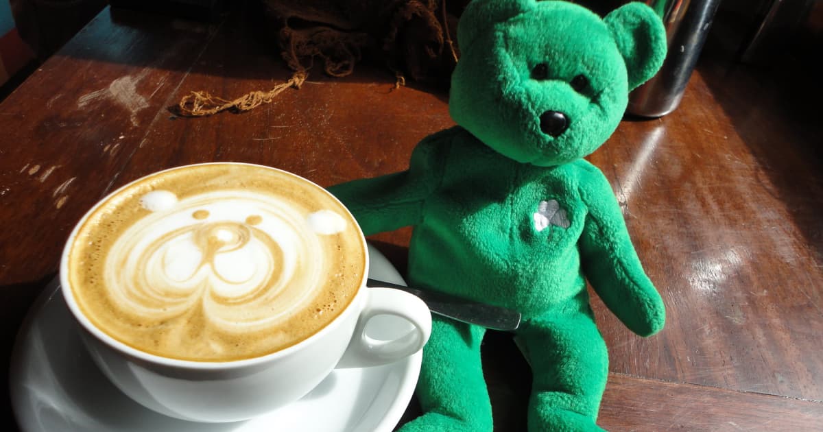 a cappuccino beside a green teddy with the teddy bears face replicated in the cappuccino foam