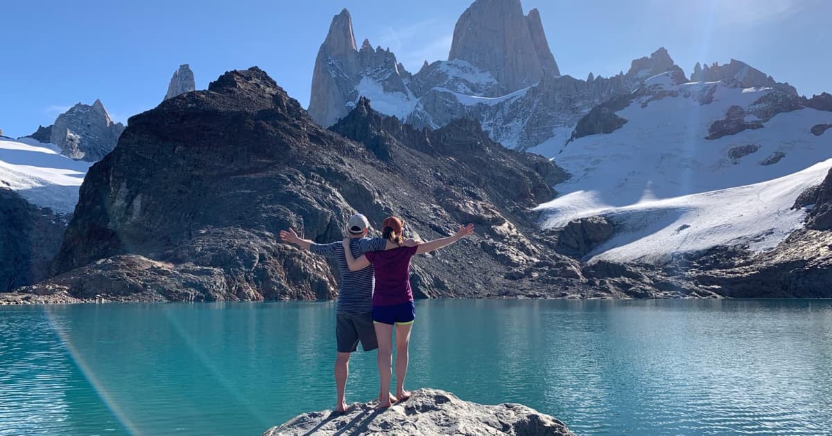 2 travelers arm in arm standing on a rock overlooking a small lake with snow capped mountains in the distance