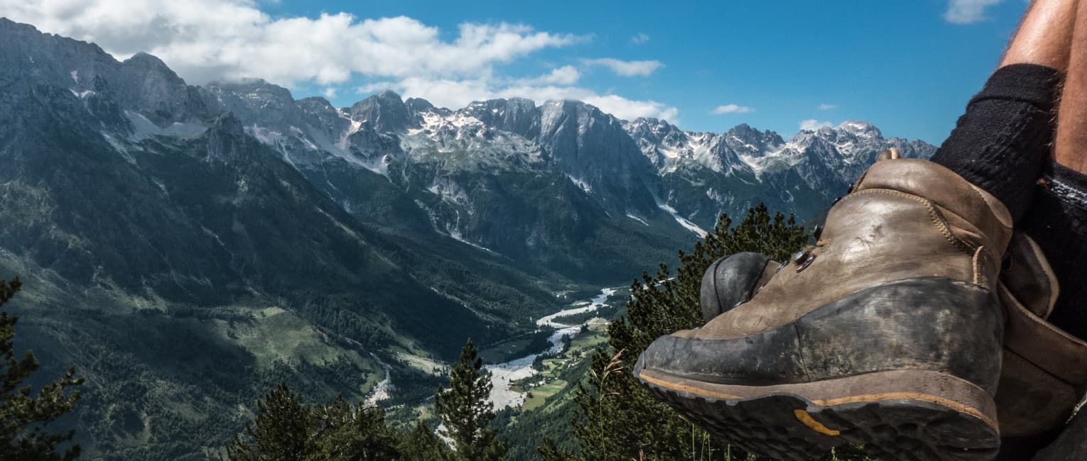 A close up of a hikers boot in front of a mountain landscape on a sunny afternoon with a blue sky