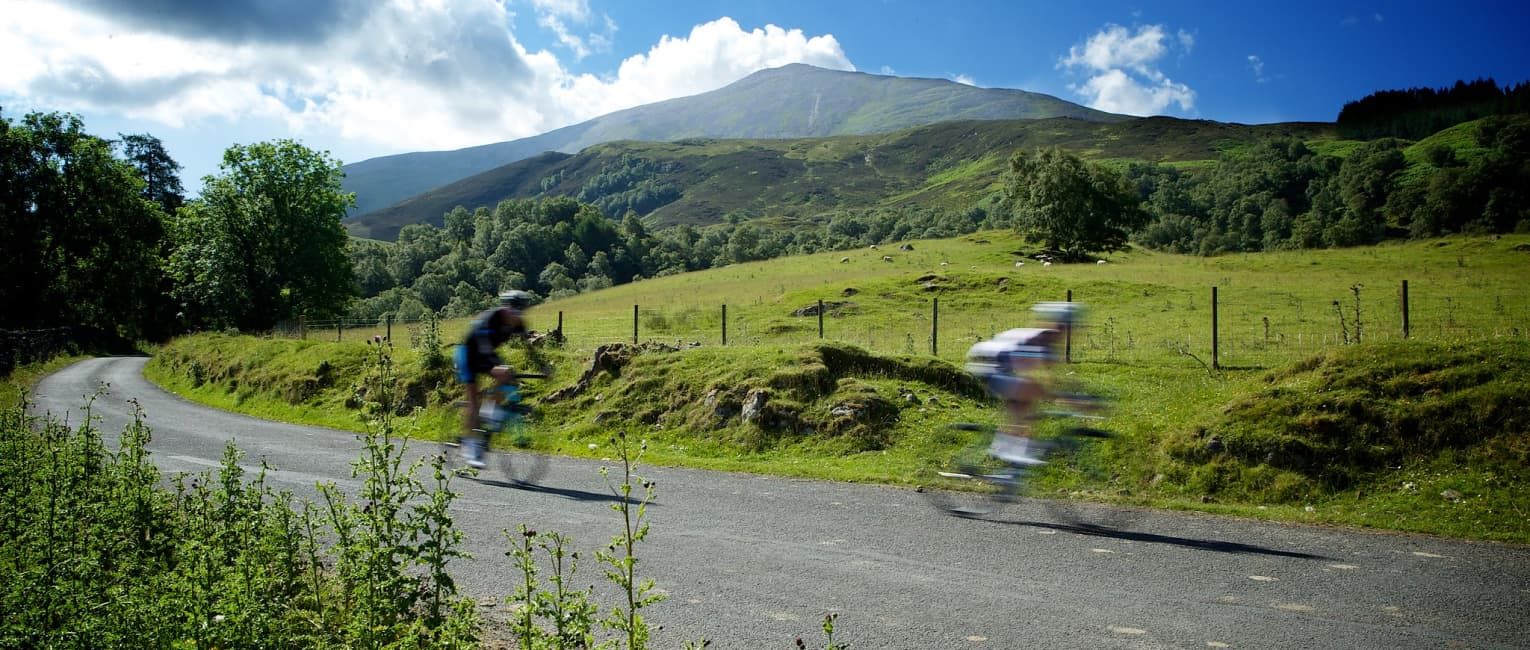 2 cyclists travelling down a hill at speed surrounded by green landscapes